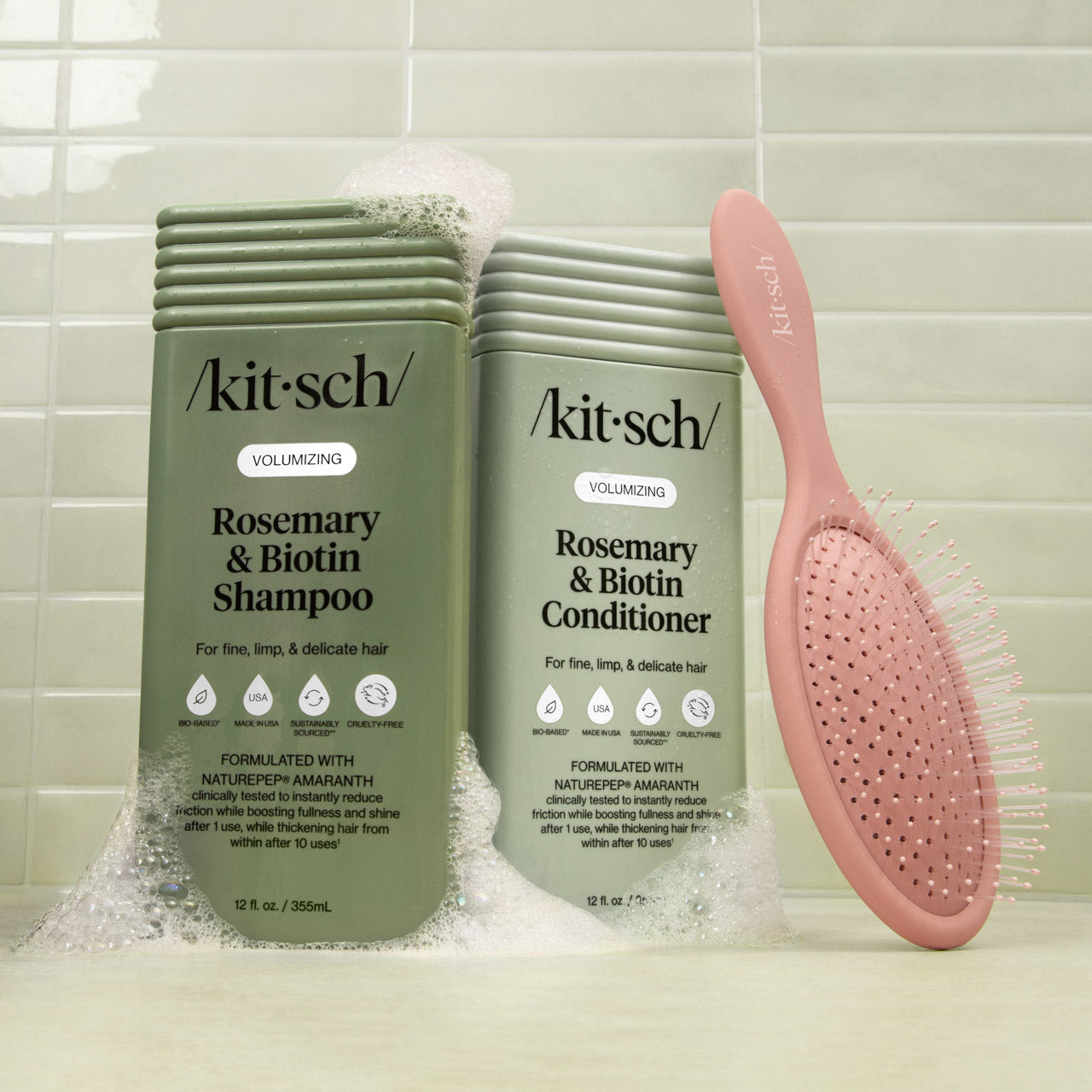 Kitsch Rosemary & Biotin shampoo and conditioner bottles with a pink brush on a tiled bathroom background.