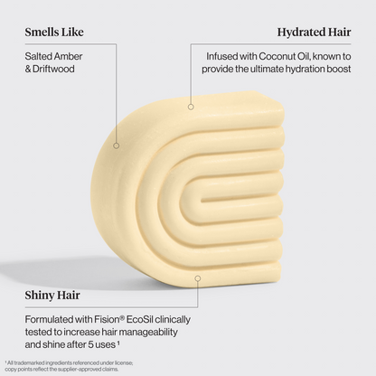 Cream-colored coconut oil conditioner bar with text describing its properties on a light gray background. Properties include: Smells like salted amber & driftwood, shiny hair -formulated with fision ecosil clinically tested to increase hair manageability and shine after 5 uses, hydrated hair - infused with coconut oil, known to provide the ultimate hydration boost